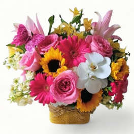 Bright mixed bouquet of pink roses, gerbera daisies, sunflowers, and white orchids in a basket with a pink butterfly.