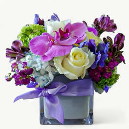 Compact arrangement of pink orchids, white roses, and mixed purple flowers in a cube vase with lavender ribbon