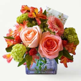 Peach roses, pink gerbera daisies, and orange alstroemeria in a purple glass cube vase.
