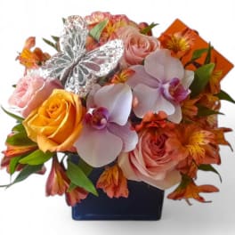 Compact arrangement of roses, orchids, and alstroemeria in a blue cube vase with a silver butterfly decoration
