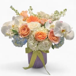 Peach and white mixed flower arrangement in a lavender vase with green ribbon