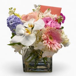 Square glass vase with pink, white, and lavender flowers and a small coral card tucked in back.