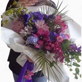 Large hand-tied bouquet of pink and purple flowers wrapped in white paper with a purple ribbon