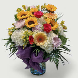 Mixed arrangement of sunflowers, roses, and daisies in a blue glass vase with a purple ribbon
