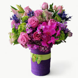 Bright pink and purple flower arrangement in a purple-wrapped vase with green ribbon and butterfly