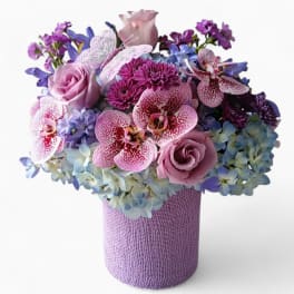 Lavender, pink, and blue flower arrangement with orchids and roses in a textured lavender cylinder vase