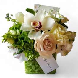 Small cube arrangement of white orchids, blush roses, and green blooms in a green vase with a card.
