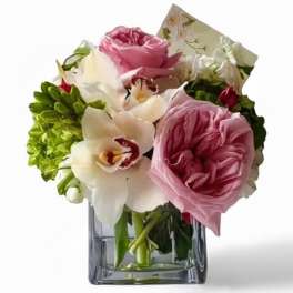 Compact arrangement of pink roses and white orchids in a clear square glass vase with a card.