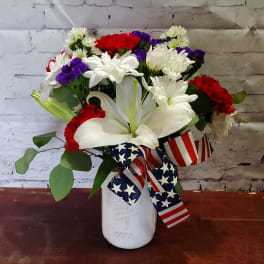 Bouquet of white lilies, red carnations, and purple daisies in a white vase with patriotic ribbon