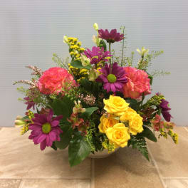 Mixed bouquet of yellow roses, purple daisies, and pink carnations in a white bowl vase