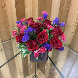 Red roses, pink carnations, and purple flowers arranged in a dark red cube vase