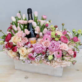 Rectangular pink and peach flower arrangement with tulips, roses, and peonies surrounding a champagne bottle.