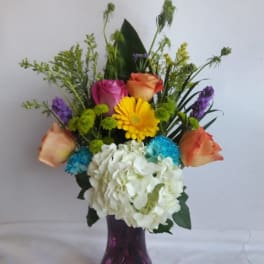 Tall mixed arrangement with roses, gerbera, hydrangea and mums in a purple glass vase