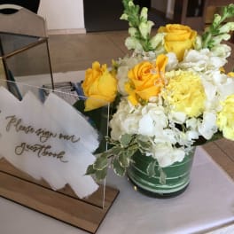 Yellow and white flower arrangement with roses and snapdragons in a green vase beside an acrylic guestbook sign