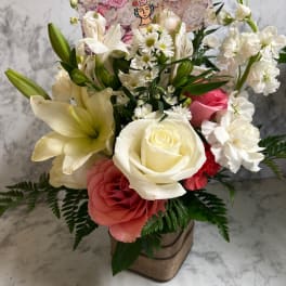 Mixed bouquet of white lilies, roses, and pink blooms in a burlap-wrapped cube vase with a floral card.