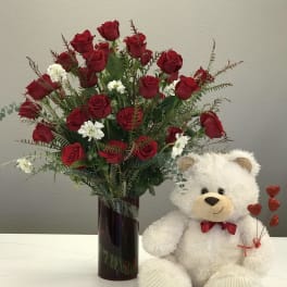Red roses in a tall vase beside a white teddy bear holding heart picks