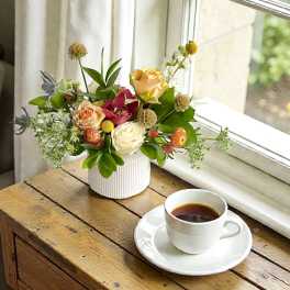 Mixed flower arrangement in a white vase beside a cup of coffee