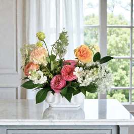 Low white bowl of pink, peach, and yellow flowers with white accents on a marble countertop.