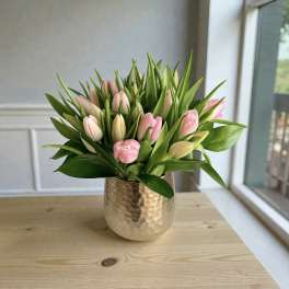 Pink and cream tulips arranged densely in a hammered gold vase on a wooden table by a window