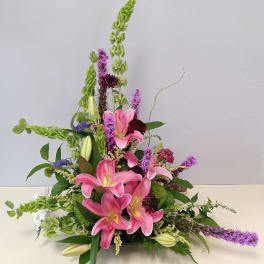 Pink lilies arranged with tall green and purple flowers in a low container