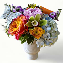 Bright mixed flower arrangement with roses, hydrangea, orchid, and butterfly pick in a white vase