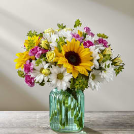 Bouquet of sunflowers, white daisies, pink carnations, and yellow roses in a green glass vase