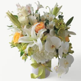 White lilies, orchids, hydrangea and roses in a white cylinder vase with a green ribbon bow.