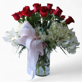 Tall glass vase arrangement with red roses, white hydrangeas, and white orchids accented by a pink ribbon bow