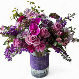Purple and pink mixed flower arrangement with roses and orchids in a clear vase wrapped with purple fabric