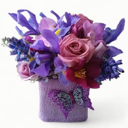 Compact purple and pink arrangement of roses, irises, orchids, and tulips in a square vase with a butterfly decoration