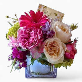 Compact bouquet of pink and cream flowers in a square glass vase with a small card.