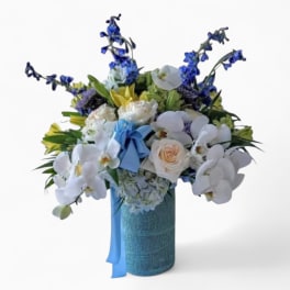 Tall bouquet of white orchids, cream roses, and blue flowers in a blue vase with a blue bow