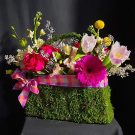 Bright mixed flowers in a moss-covered handbag container with a plaid ribbon bow