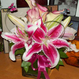 Pink and white lilies arranged in a glass vase with a ribbon and butterfly picks.
