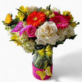 Bright mixed bouquet of white roses, gerbera daisies, and yellow blooms in a glass vase with butterfly accents