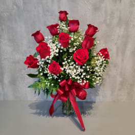 Red roses arranged in a clear glass vase with white baby's breath and a red ribbon.