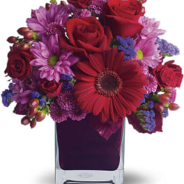 Red roses and purple daisies arranged in a square glass vase