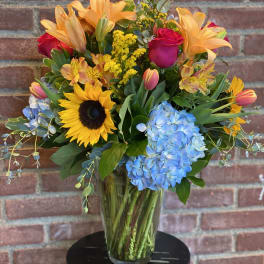 Mixed bouquet with sunflowers, roses, lilies, and blue hydrangeas in a glass vase