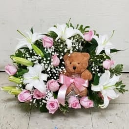 Pink roses and white lilies arranged around a teddy bear with a pink ribbon