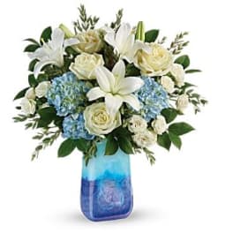White lilies and roses arranged with blue hydrangeas in a blue glass vase.