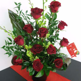 Tall arrangement of dark red roses with green orchids in a black vase and a red Happy Birthday pick