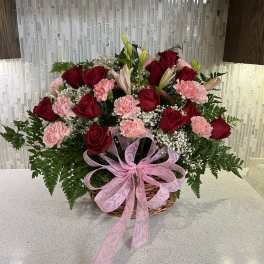 Basket arrangement of red roses, pink carnations, and lilies with a pink ribbon