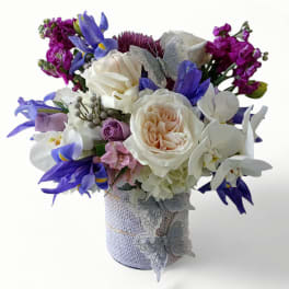 Compact arrangement of white roses, blue irises, and orchids in a wrapped vase with silver butterflies.