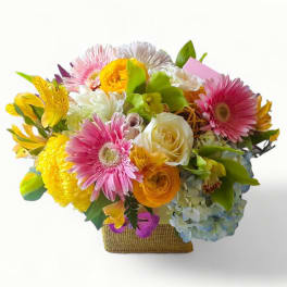 Bright mixed bouquet with pink gerbera daisies, yellow blooms, roses and hydrangea in a square burlap-wrapped vase