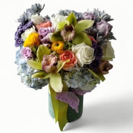Tall mixed bouquet with orchids, roses, ranunculus, and hydrangeas in a green vase with a butterfly accent.