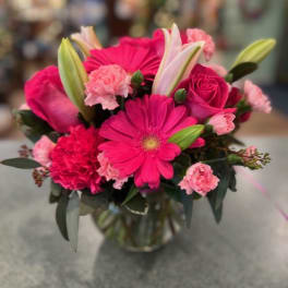 Compact bouquet of bright pink gerbera daisies, roses, carnations, and lilies in a clear glass vase