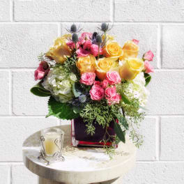 Bouquet of yellow and pink roses in a square vase with a candle beside it