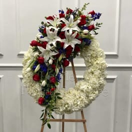 Large floral wreath on an easel with red roses, white lilies, and blue accents