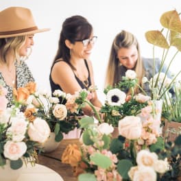 Three women arranging blush and white flower centerpieces on a wooden table