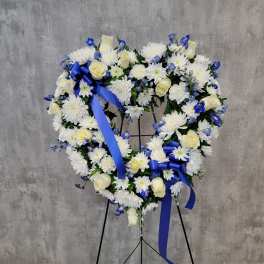 Heart-shaped floral wreath with white flowers, cream roses, and blue ribbons on a stand